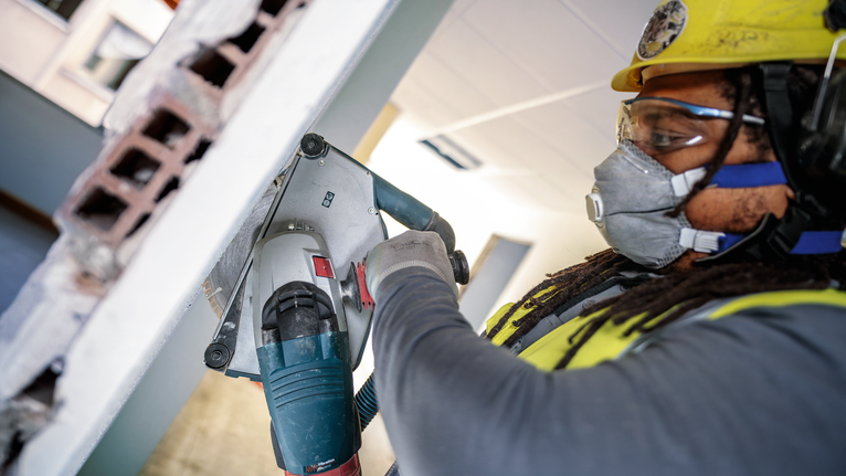 Person wearing safety equipment cuts through a wall using a power tool.