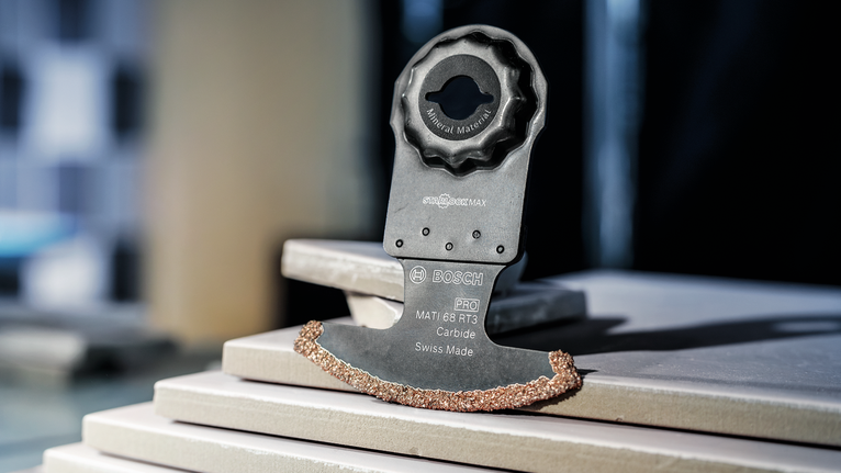 PRO MATI 68 RT3 Multitool Blade, 2.8 x 68 mm Oscillating tool blade with carbide edge rests on stacked ceramic tiles.