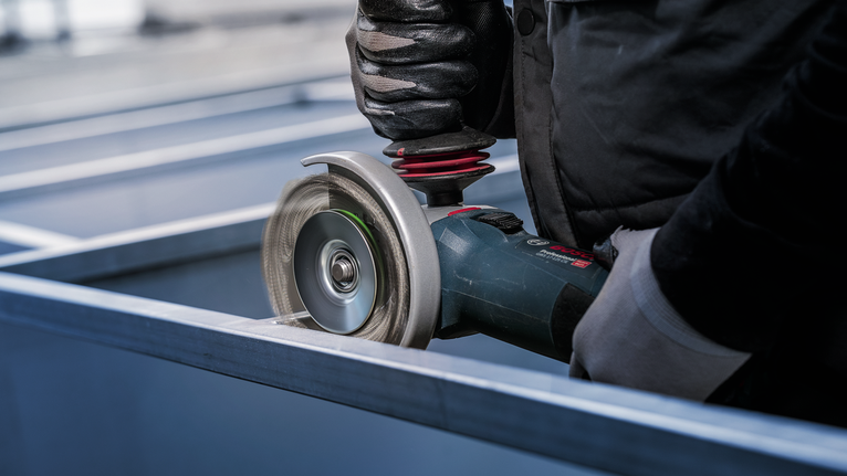 PRO Metal clean Bevel Brush, 100 x 0.3 mm, Thread Person wearing safety equipment grinds a metal beam with an angle grinder.