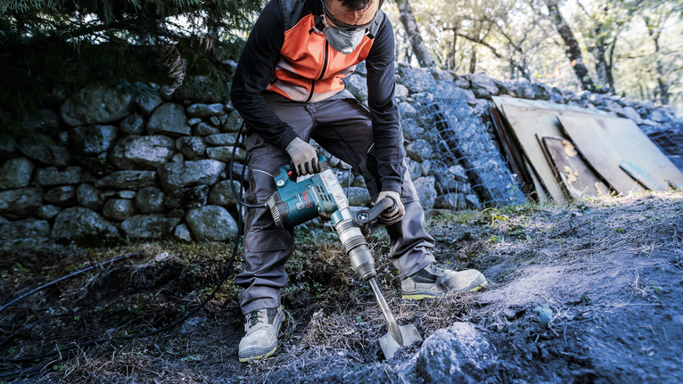 PRO SDS max Excavation Chisel, 110 x 400 mm Person wearing safety equipment uses a power tool to break rocky ground outdoors.