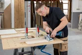 A person wearing safety equipment uses a biscuit joiner on clamped wood pieces.