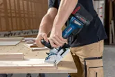 A person wearing safety equipment uses a biscuit joiner to cut wood on a workbench.