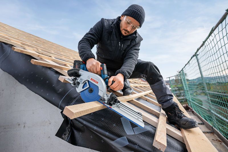 A person wearing safety equipment cuts roof beams with a circular saw on a sloped structure.