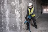A person wearing safety equipment uses a cordless demolition hammer on a concrete wall.