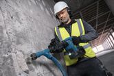 A person wearing safety equipment uses a cordless demolition hammer to break concrete.