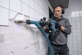 A person wearing safety equipment uses a cordless demolition hammer to break wall tiles.