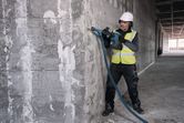 A person wearing safety equipment uses a cordless demolition hammer on a concrete wall.