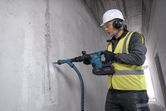 A person wearing safety equipment uses a cordless demolition hammer on a concrete wall.