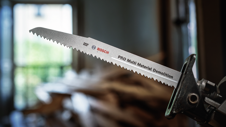 PRO Multi Material Demolition S1110VF Blade, 1.6 x 22 x 225 mm, 5 pcs Reciprocating saw blade attached to a power tool, background shows blurred wood debris.