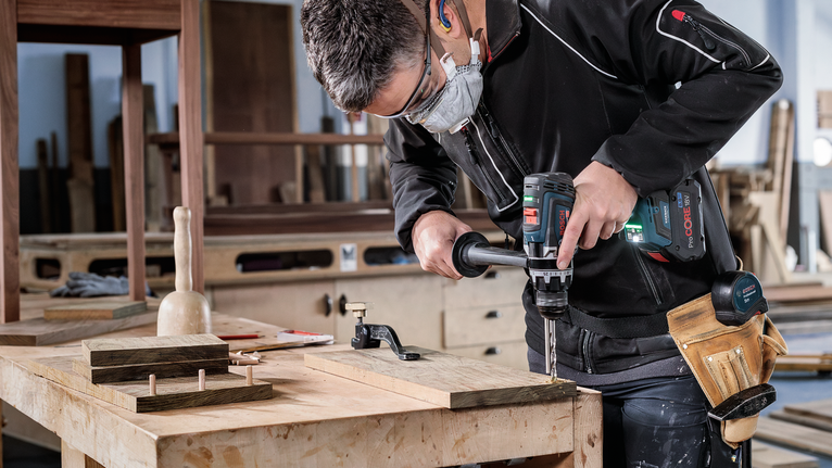 Person wearing safety equipment drills into wood at a workbench.
