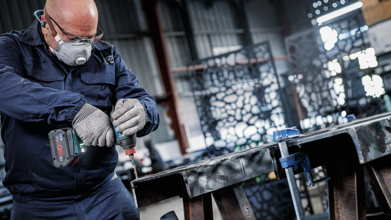 A person wearing safety equipment drills into a metal beam in an industrial workshop.