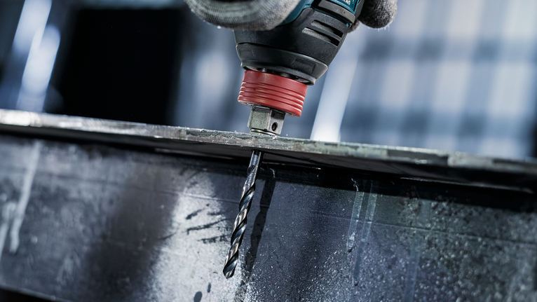 Person wearing safety equipment drills a hole into a metal beam.