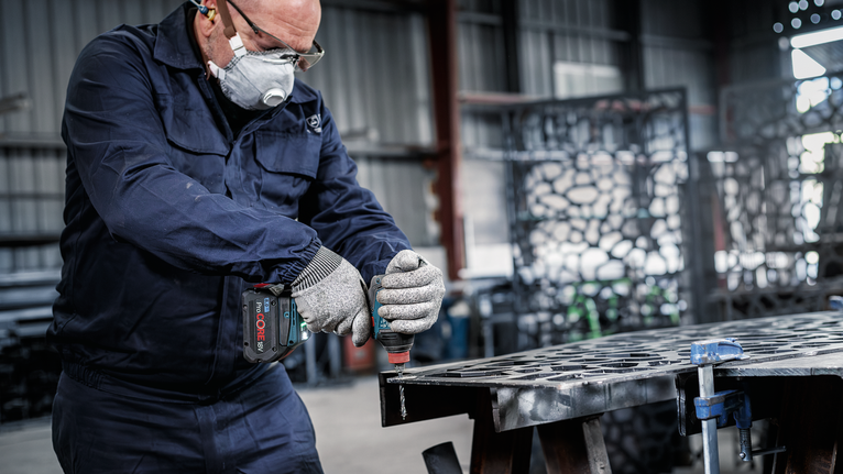 Person wearing safety equipment drills into a metal sheet in a workshop.