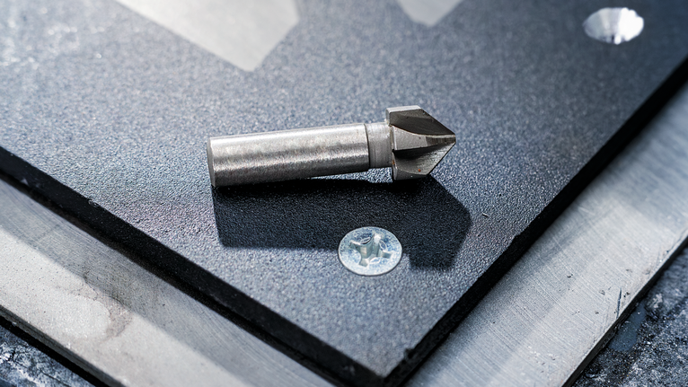 PRO Steel HSS Countersink Bit, 12 x 40 mm, M6 Countersink drill bit resting on a metal sheet near a screw and drilled holes.