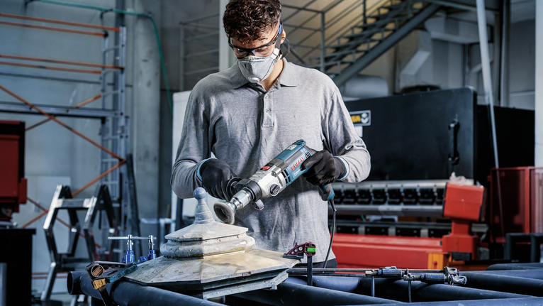 PRO Metal clean Cup Brush, Straight Shank, 50 x 0.2 mm Person wearing safety equipment drills into a metal fixture in an industrial workshop.