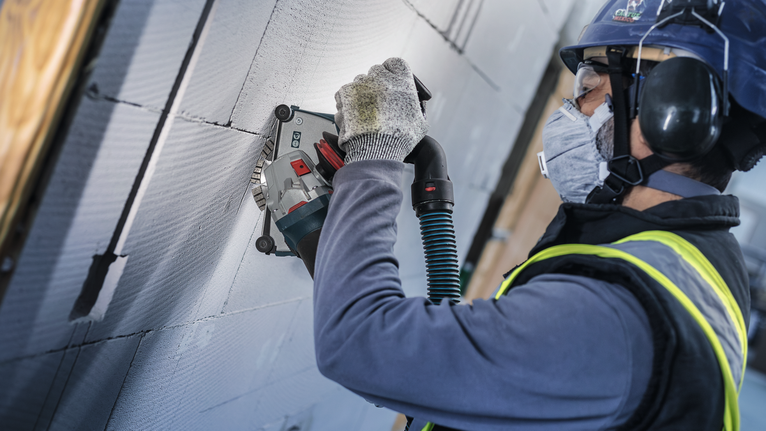 Person wearing safety equipment grinds a groove into a concrete wall using a power tool.