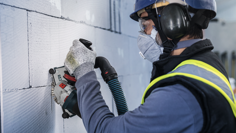 Person wearing safety equipment uses a power tool to cut grooves into a concrete wall.