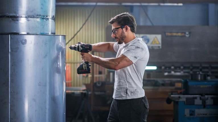 Person wearing safety equipment drills into a large metal cylinder in a workshop.