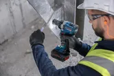 A person wearing safety equipment uses a cordless impact wrench on a metal frame.