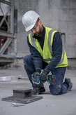 A person wearing safety equipment uses a cordless impact wrench on a construction site.