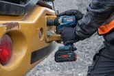 A person wearing safety equipment uses a cordless impact wrench on construction machinery.