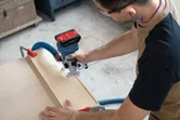 A person wearing safety equipment uses a cordless router to shape wood on a workbench.
