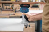 A person guides a cordless router along the edge of a white board.