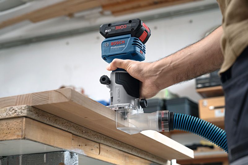 A person uses a handheld router with dust extraction on the edge of a wooden board.