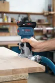 A person trims the edge of a wooden board with a cordless power tool attached to a dust hose.