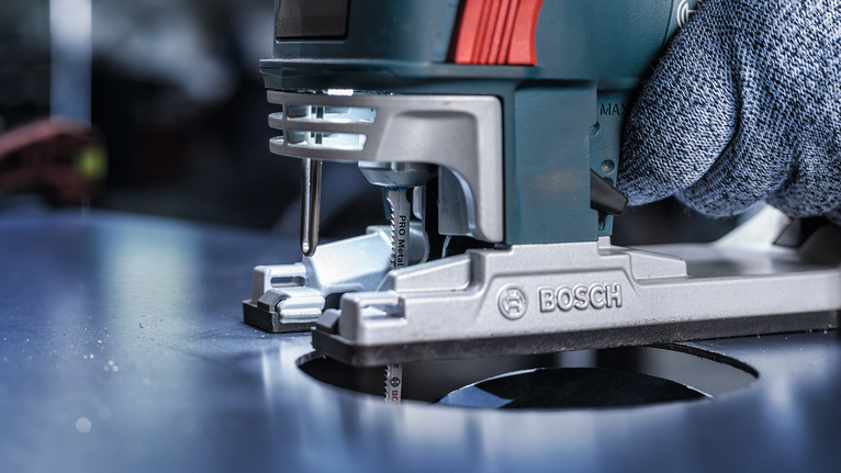 Person wearing safety equipment guides a jigsaw to cut a circular hole in metal.