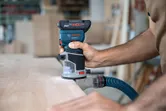 A person guides a cordless palm router along the edge of a wooden board.