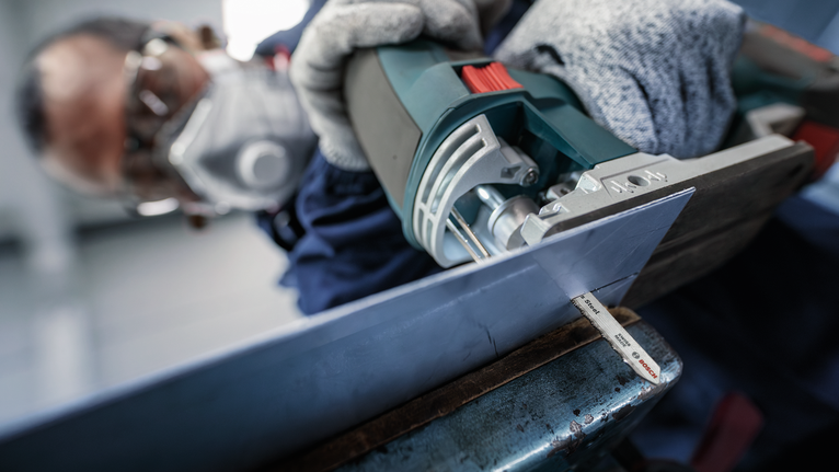 Person wearing safety equipment uses a jigsaw to cut a metal sheet.