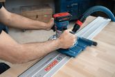 A person guides a cordless palm router along a track on a wooden workbench.