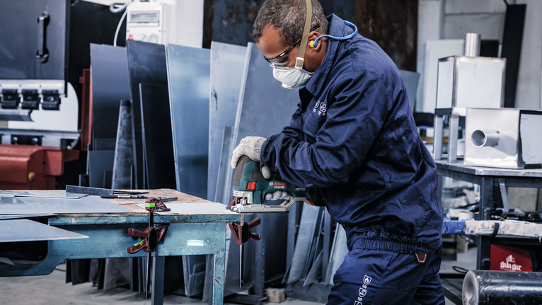 Person wearing safety equipment cuts a metal sheet with a power saw in a workshop.