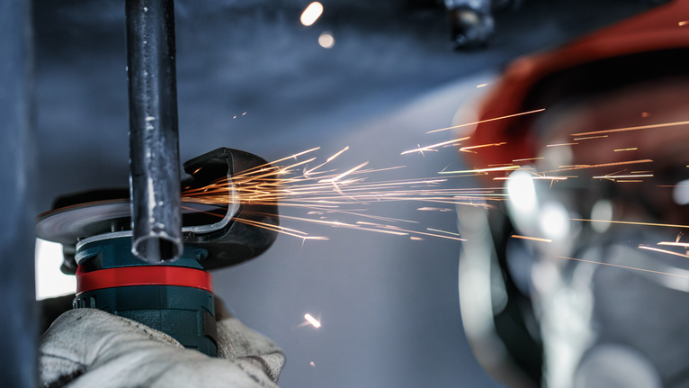 Worker wearing safety equipment grinds metal pipe, creating sparks.