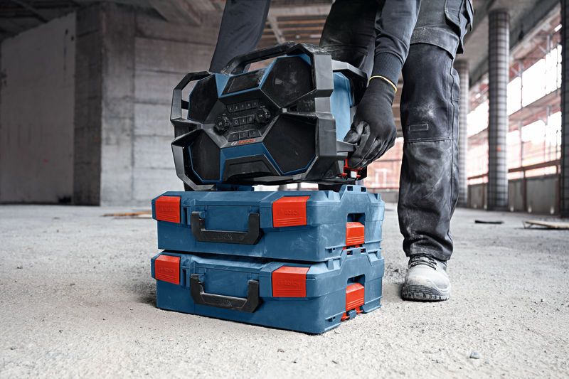 A person wearing safety equipment stacks a rugged jobsite radio on toolboxes in a construction area.