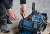 Person wearing safety equipment plugs a charging cable into a rugged jobsite radio.