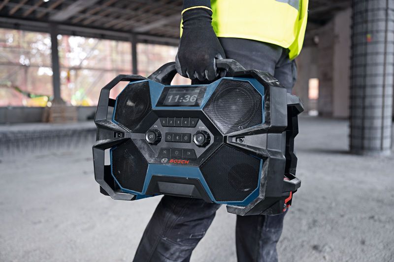 A person wearing safety equipment carries a rugged jobsite radio inside a construction area.