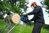 A person wearing safety equipment uses a chainsaw to cut a log outdoors.