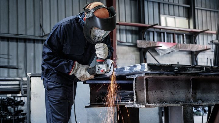 A worker wearing safety equipment grinds a metal beam with sparks flying.