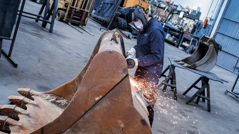 A person wearing safety equipment grinds a large excavator bucket, producing sparks.