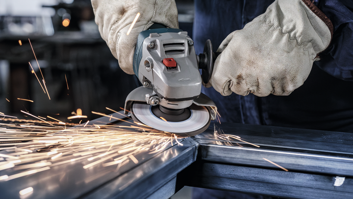 Person wearing safety equipment uses an angle grinder to cut metal, producing sparks.
