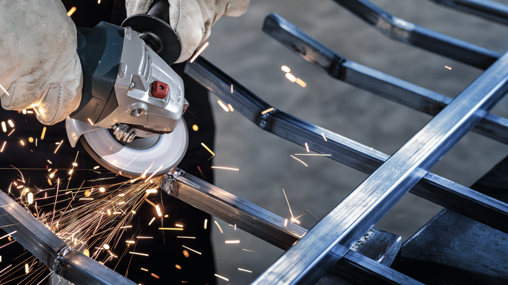 A worker wearing safety equipment grinds a metal frame, producing sparks.