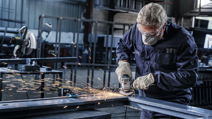 A worker wearing safety equipment grinds a metal beam, creating bright sparks.