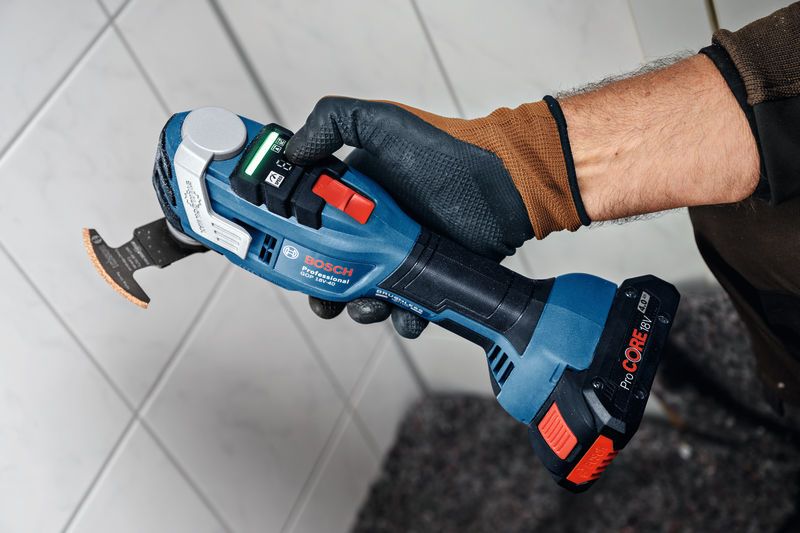 A person wearing safety equipment holds a cordless multi-cutter near a tiled wall.