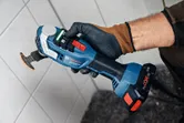 A person wearing safety equipment holds a cordless multi-cutter near a tiled wall.