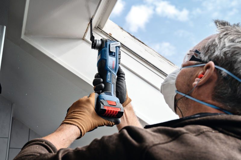 A person wearing safety equipment uses a cordless multi-cutter to trim a window frame.