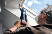 A person wearing safety equipment uses a cordless multi-cutter to trim a window frame.
