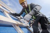 A person wearing safety equipment uses a cordless drill to fasten wood on a sloped surface.