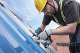 A person wearing safety equipment uses a cordless power tool to fasten screws on a metal roof.
